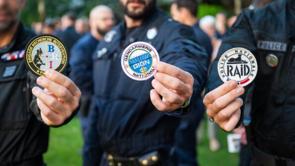 Trois hommes floutés tendant vers l'objectif un écusson, représentant leur unité d'appartenance : BRI, GIGN, RAID. Ils se trouvent dans un jardin. Derrière eux, on aperçoit de nombreuses personnes, également floutées.