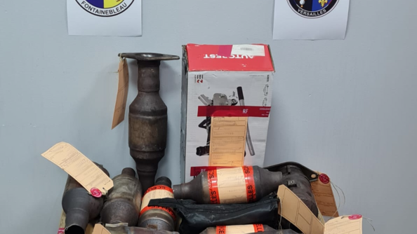 des pots catalytiques avec des cartons de scellés contre un mur gris. Sur le mur les rondaches de la section de recherches de Versailles et la brigade de recherches de Fontainebleau