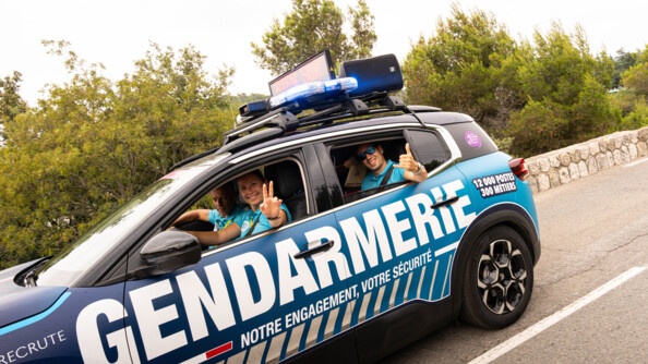 Une voiture de couleur bleue marine de la gendarmerie roule sur la route. On aperçoit les gendarmes qui sont à son bord, souriants, et effectuant le signe de la victoire avec leurs doigts, par les fenêtres ouvertes.