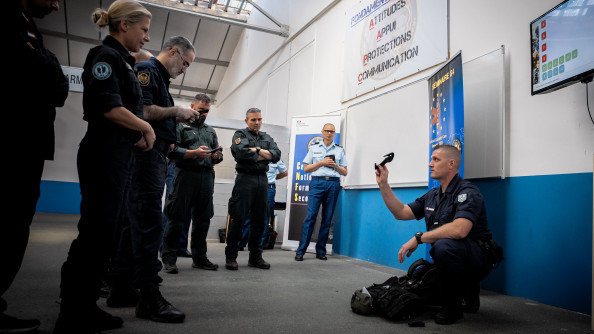 Un gendarme accroupi par terre montre un outil à d'autres personnes debout. La photo est prise en intérieur et de jour.
