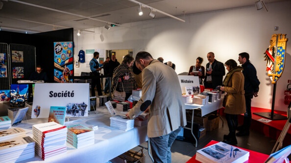 Salon littéraire et artistique avec des livres posés sur 2 tables correspondant à 2 thèmes : la société et l'histoire. On aperçoit derrière l'emblème de la promotion Connétable Bertrand Duguesclin et des oeuvres artistiques, des peintures plus précisemment.