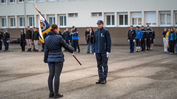 Une femme tenant un fanion de dos, devant elle un cadet de la gendarmerie au garde-à-vous. Autour, les gendarmes en rang en cérémonie
