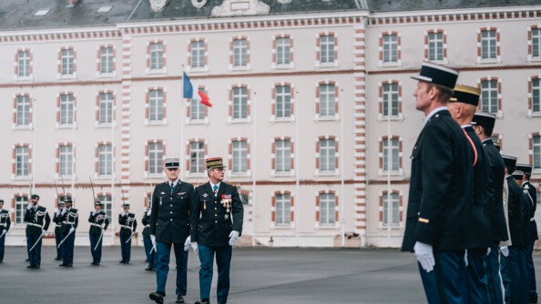 Un général de gendarmerie, suivi par un colonel, passe en revue ses troupes sur une place d'armes devant un bâtiment d'etat-major