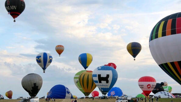 dans une prairie, les ballons se gonflent d'ai et décollent. Des visiteurs regardent.
