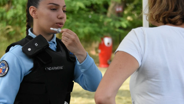 Une gendarme de face fait une d&eacute;monstration de l'utilisation d'un &eacute;thylotest &agrave; une personne civile en tee-shirt blanc, vue de dos.