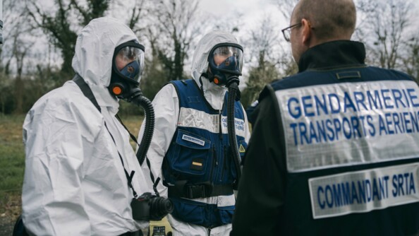 Les gendarmes TICS dans leur tenue d'intervention. Ils ont une combinaison blanche avec une capuche. Les deux ont un masque qui recouvre tout leur visage, l'un a un appareil photo autour du cou.