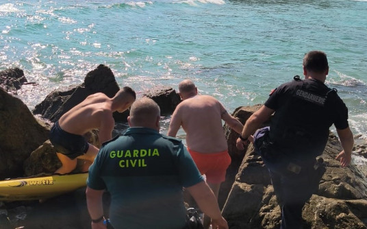 Sur une côte rocheuse, un sauveteur des plages, un agent de la Guardia Civil et un gendarme français aident un homme en short orange à sortir de l’eau. Un kayak jaune est visible, la mer est calme et brille sous le soleil..