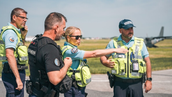 Quatre gendarmes, dont trois avec une chasuble jaune de la GTA et le dernier en noir. Celle du milieu, une femme, montre du bras quelque chose au loin.