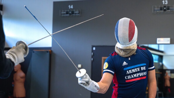 Maxime Pauty vêtu du survêtement bleu et rouge de l'armée de champions s'entraîne face à son maître d'arme dont on voi seulement le fleuret à gauche