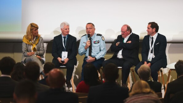 Au premier plan flou, toris rangs de personnes assistant à la table ronde. Au deuxième plan, de gauche à droite sur des tabourets hauts: Dominique Faure, un élu, le DGGN (le général d'Armée Christian Rodriguez) s'exprimant au micro, et deux autres élus.