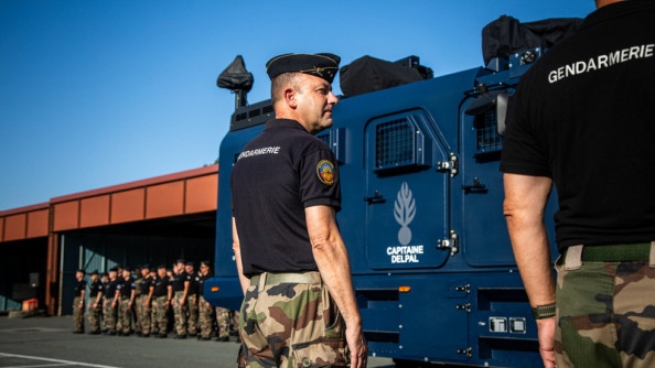 La photo représente deux militaires, debout, de dos, au passage d'un véhicule blindé de la gendarmerie. En face d'eux se trouve un groupe de militaires.
