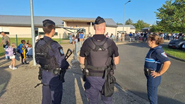 Deux gendarmes de dos, avec à droite de l'iùage une femme de la police municipale de profil, aux abords d'une école. A droite, la route