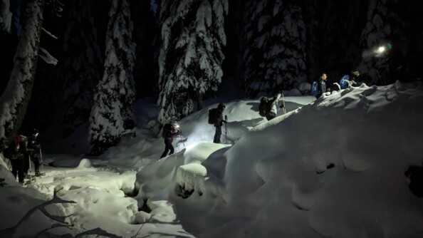 Des marcheurs progressent les uns derrière les autres sur un sentier enneigé, bordé de sapins. Il fait nuit. Tous sont équipés d'une lampe frontale et de batons de marche