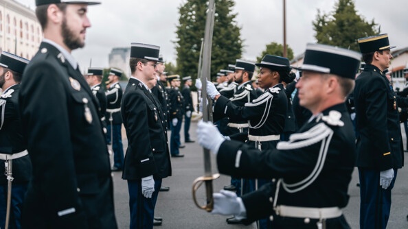 Plusieurs rangs de gendarmes en tenue de cérémonie se font face, ceux du rang de droite sont en train de tenir la sabre par le manche et la lame