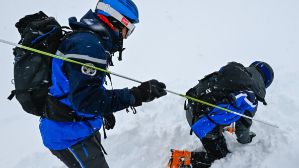 Deux gendarmes l'un deriière l'atre dans la neige, l'un debout avec une perche métallique, le premier un genou en terre