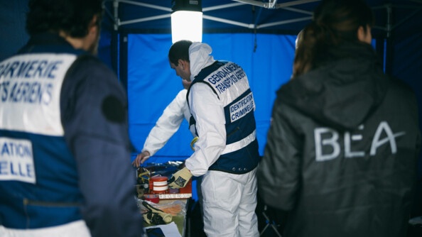 De dos, à gauche un gendarme des transports aériens, à droite, un membre du BEA. Au centre, les deux gendarmes TIC qui déposent les éléments récupérés.