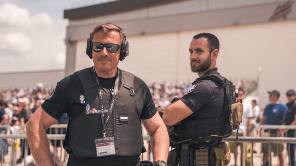 Devant une barrière délimitant la zone public du Bourget, un gendarme de la GTA vêtu de noir, un casque anti-bruit sur les oreilles, les mains sur les hanches. A droite, de profil, un de ses collègues, le regard dans notre direction