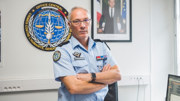 Le général Jean-Philippe Reiland, debout, les bras croisés, en tenue de gendarmerie, chemisette bleue. Derrière lui, sur le mur, la reproduction de la rondache de l'OCLCH et le portrait du président de la République.