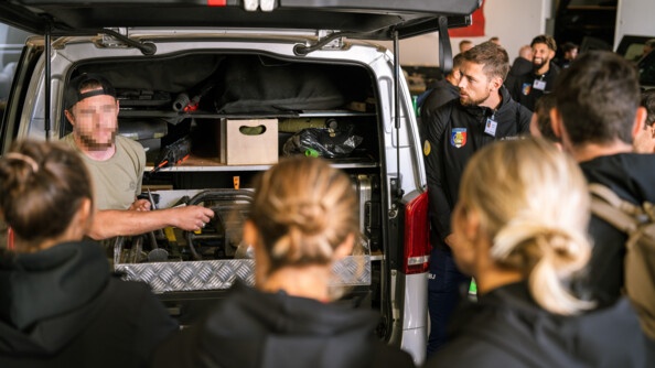 Devant un véhicule dont le coffre est amenagé pour du matériel, un gendarme du GIGN montre le matériel utilisé. Il est entouré par les sportifs