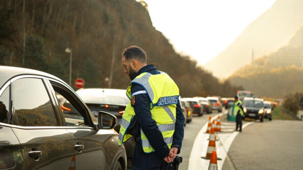 Un gendarme avec une chasuble jaune conseille un automobiliste sur la route. On peut voir une longue colonne de véhicules.