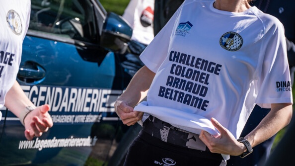 Photo d'une femme dont on aperçoit uniquement les bras et le torse, vêtue d'un tshirt blanc portant la mention "Challenge colonel Arnaud Beltrame".  Juste derrière elle, on aperçoit la portière d'un véhicule bleu de la gendarmerie nationale.