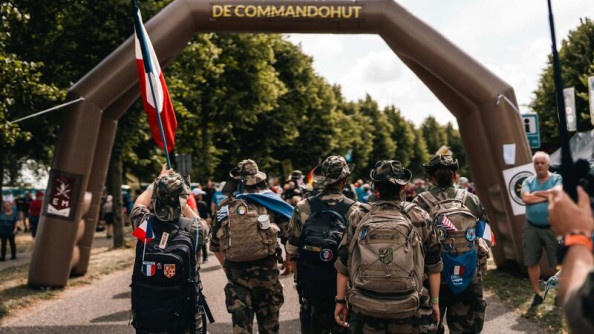 Un groupe de militaires, en tenue kakie, avance, de dos, s'apprêtant à passer sous un auvent gonflable de couleur marron. L'un des militaires porte un drapeau français.