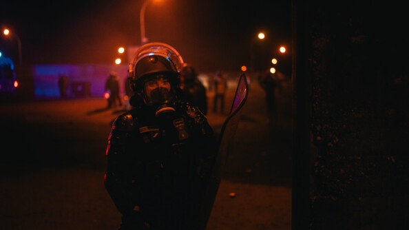 Une femme gendarme, portant une tenue noire, un casque et un masque, se tient debout, face à l'objectif. La scène se déroule de nuit.