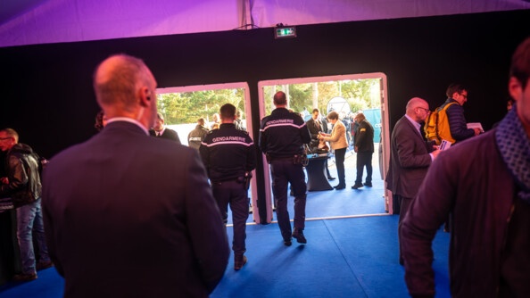 Dans un espace intérieur donnant sur l'extérieur par deux ouvertures, on apercoit de dos au premier plan un holle en tenue civile, équipé d'une oreillette. Devant lui, au niveau des ouvertures, se tiennent de dos deux gendarmes. Au dos de leur veste bleue marine, est inscrite la mention Gendarmerie. Au sol, une moquette bleue. Et une lumière rose au niveau du plafond. Un point d'accueil, où se pressent des visiteurs, est installé à l'extérieur