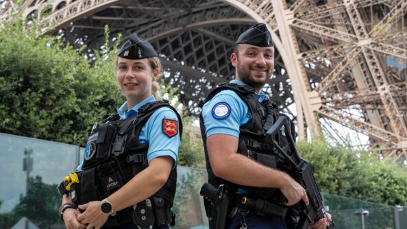 Deux jeunes gendarmes  - un homme et une femme - se tiennent côte à côte, devant la Tour Eiffel. Ils sont équipés d'un gilet pare-balles. Le gendarme situé à droite tient dans ses mains un fusil.