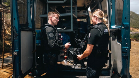 Deux gendarmes mobiles, un homme à gauche et une jeune femme à droite, vus de trois-quarts dos, se font face devant les portes arrière ouvertes d'un Irisbus.