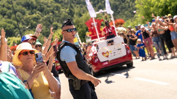 Un gendarme se tient debout, au milieu de la foule, le sifflet dans la bouche, équipé d'un gilet pare-balles et de lunettes de soleil, alors que passent sur la route les véhicules de la caravane du Tour de France 2024. Le temps est ensoleillé et les nombreux spectacteurs présents sur le bord de la route portent pour la plupart un chapeau.