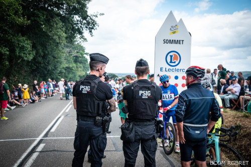 Deux gendarmes d'un Peloton de surveillance et d'intervention de gendarmerie (PSIG), vus de dos, sont au bord de la route. Ils échangent avec un cycliste amateur, casque sur la tête et vélo à la main, vu de dos. Au bord de la route, de part et d'autre de la chaussée, sont massés de nombreux spectateurs, debout et assis.