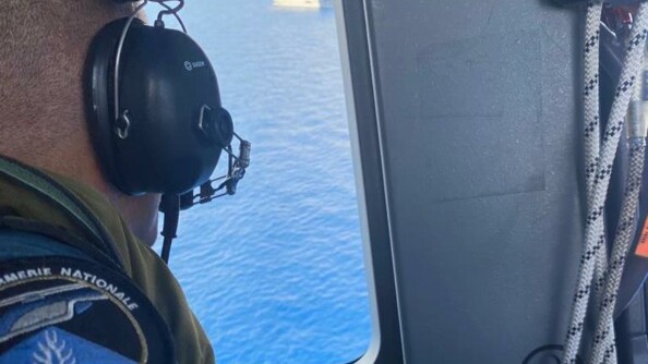 Vue sur la mer et le porte hélicoptères Mistral depuis le hublot d'un hélicoptère de la gendarmerie nationale. Au premier plan un militaire de l'équipage.