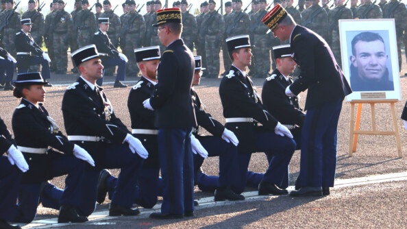 En arrière plan, des élèves-gendarmes en treillis, famas en main, au garde-à-vous.  Devant eux, une première rangée d'élèves-gendarmes en vareuse, agenouillés. Au premier plan, une rangée de sept élèves-gendarmes, en vareuse, agenouillés, auxquels le major général et le commandant de région, de dos, remettent successivement les galons. A droite, un portrait, sur chevalet, du parrain de promotion.