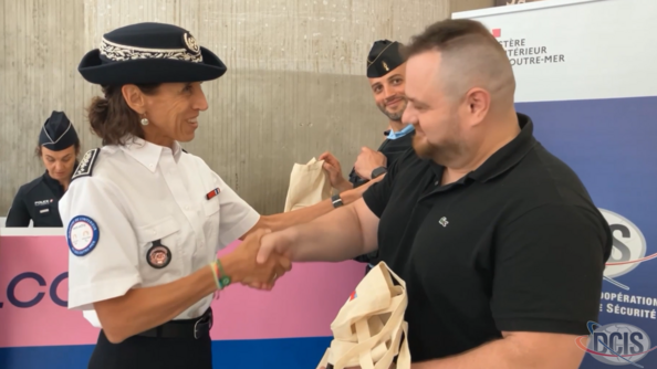 Une femme, membre des forces de l'ordre, serre la main d'un citoyen, un homme d'âge moyen vêtu d'un polo noir. Derrière eux se dresse un kakémono du Ministère de l'Intérieur et des Outre-mer, portant également la mention DCIS