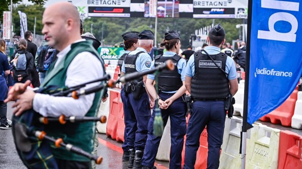 Au premier plan à gauche, flou, un joueur de cornemuse à la veste verte en train de parler vers la gauche. Au milieu, quatre gendarmes de dos devant le barrièrage plastique rouge et blanc. A droite un drapeau bleu de radio France. Autour ,du public, desarcades siglées Bretagne
