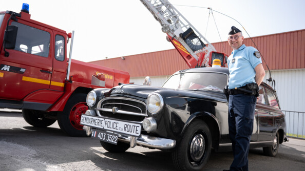 Un gendarme debout et souriant, les bras derrière le dos, devant une vieille peugeot 403 noire. A gauche un vieux véhicule de pompiers rouge