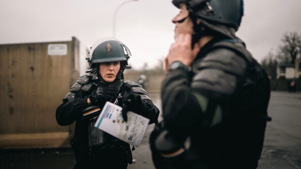 Une femme gendarme revêtant un uniforme et des gants noirs et un casque se tient debout face à un homme apparaissant de biais sur la photo. La femme tient un papier dans sa main gauche, sur lequel figure un plan.