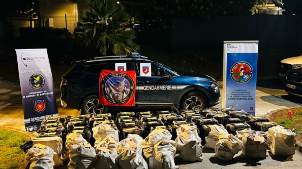 Photo de nuit montrant une voiture de gendarmerie devant laquelle plusieurs dizaines de sacs beiges sont posés. De chaque côtés, deux kakemono, un de la S.R de Saint-Martin ( à gauche) et un du COMGENDSBSM, sont positionnés.