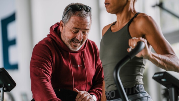 Une sportive vêtue d'une tenue grise, effectue un exercice sur un appareil dans une salle d'entraînement physique. Son entraîneur, qui se tient debout à ses côtés, accompagne la sportive en donnant des instructions. La photo est prise de biais.