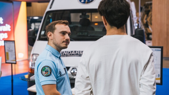 Un gendarme vêtu d'un polo bleu ciel discute un homme brun, vêtu d'un t-shirt à manches longues de couleur blanche, se tenant également debout, de dos. On aperçoit en arrière plan une camionnette blanche de la gendarmerie. La scène se déroule au salon de l'Auto 2024, à Paris.