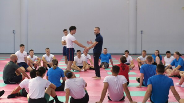 La scène se déroule sur un tatami. Les élèves sont assis en cercle autour d'un cadre et d'un élève. Le cadre montre une prise pour se défendre