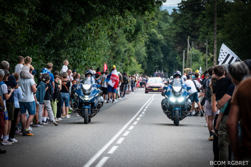 Deux motocyclistes de la gendarmerie ouvrent la route d’une course cycliste féminine, suivis de près par un véhicule de la course et un peloton de coureuses. Les motos bleues sont équipées de gyrophares allumés. De nombreux spectateurs bordent la route des deux côtés, applaudissant et filmant les cyclistes. L’ambiance est festive et animée.
