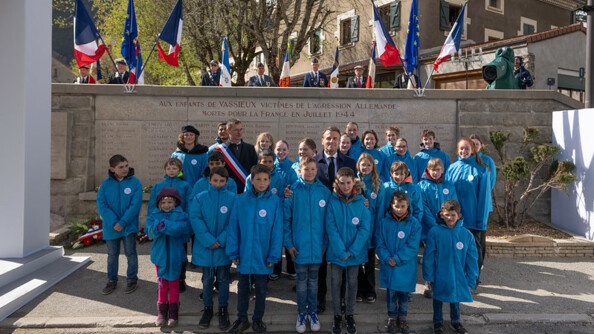 Le président de la république, Emmanuel Macron, apparait de face, debout, entouré d'enfants vêtus du même blouson bleu, devant un monument aux morts, sur lequel est inscrit "Aux enfants de Vassieux victimes de l'agression allemande morts pour la France en juillet 1944