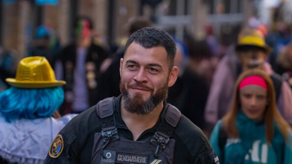 La photo repr&eacute;sente un gendarme, debout, dans la rue, entour&eacute; de carnavaleux, qui portent un d&eacute;guisement, lors du carnaval de Bergues.