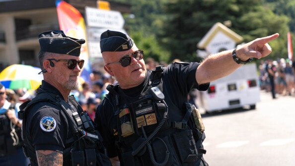 Deux gendarmes revêtant un uniforme noir, discutent ensemble sur la route. L'un désigne un point à l'horizon, en pointant l'index de sa main gauche. Derrière eux, on devine la foule, ainsi qu'un véhicule de type camionnette, évoluant sur la route. Le temps est ensoleillé.