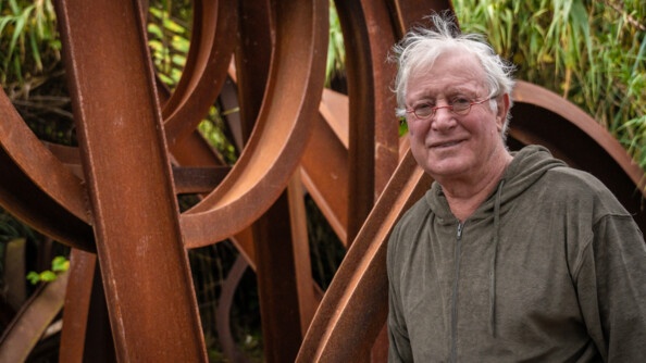 Photo du sculpteur Jean-Pierre Rives, posant devant l'une de ses oeuvres, faite de courbures en acier rouillé. L'homme aux cheveux blancs porte un gilet de couleur kakie, ainsi que de petites lunettes rouges de forme ronde.