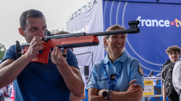 Une homme vêtu d'un t-shirt bleu, tire avec une carabine orange sur un stand de tir. A côté de lui se tient une femme gendarme qui rigole.