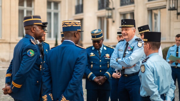 Dans une cour extérieure pavée se tiennent deux autorités de la gendarmerie nationale, entourées de gendarmes congolais. Derrière eux, se trouve un bâtiment formant un angle, dont on distingue les fenêtres