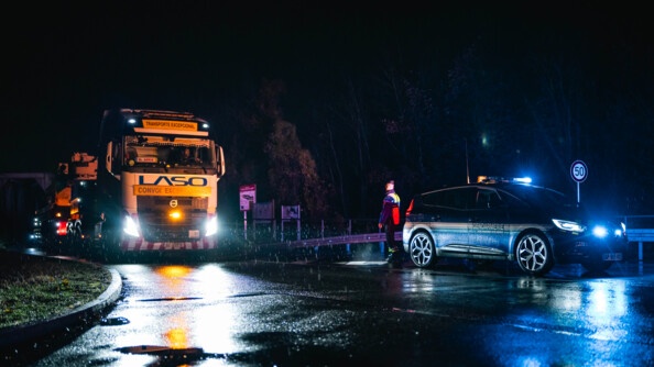 Le convoi arrive sur une petite route ou un gendarme se tient présent, devant son véhicule. Il fait nuit et il pleut.